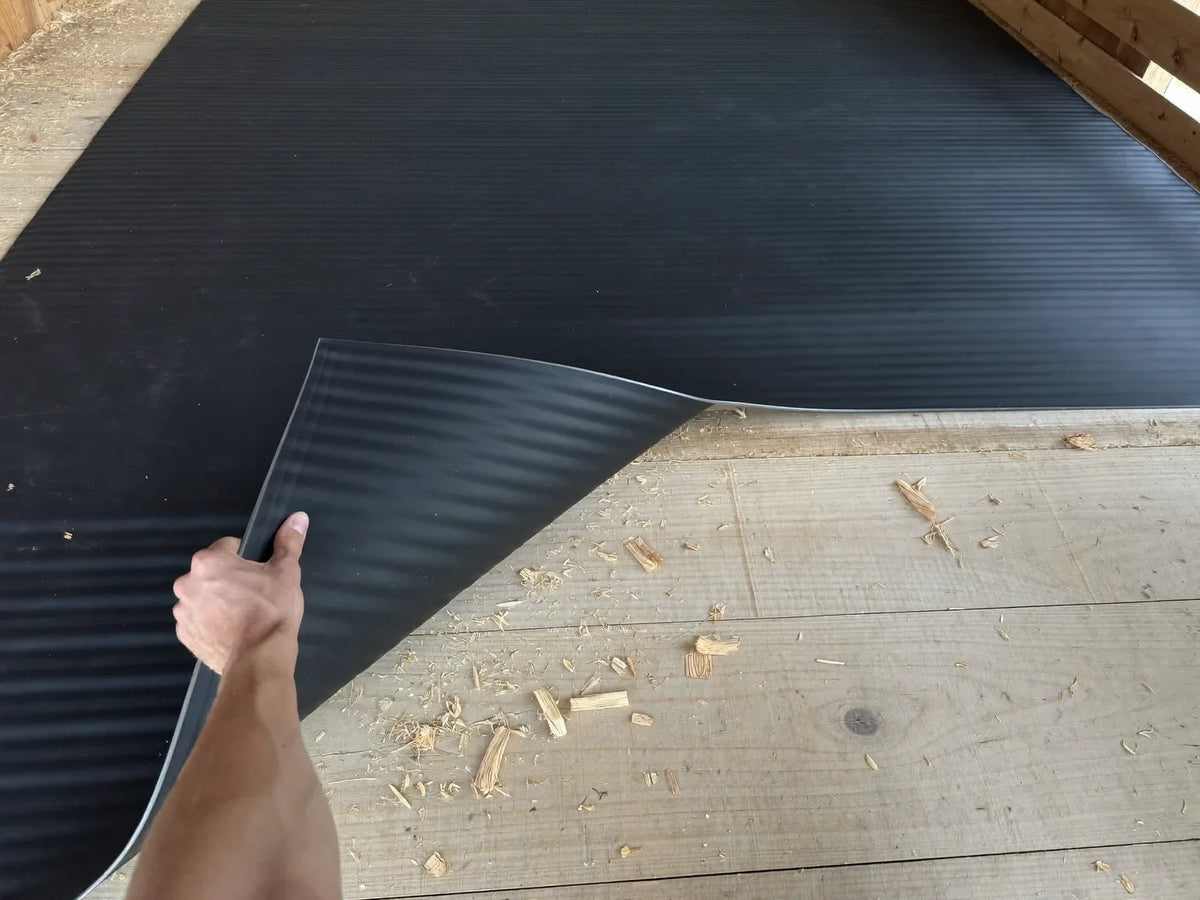 Hand lifting the corner of a black rubber mat off the bare timber floor of an empty chicken coop