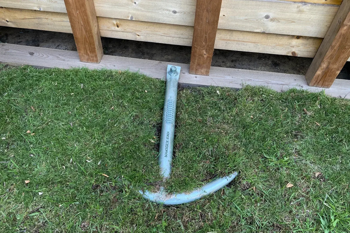 Galvanised screw-in earth anchor turned into mown grass beside the bottom rail of a timber coop frame