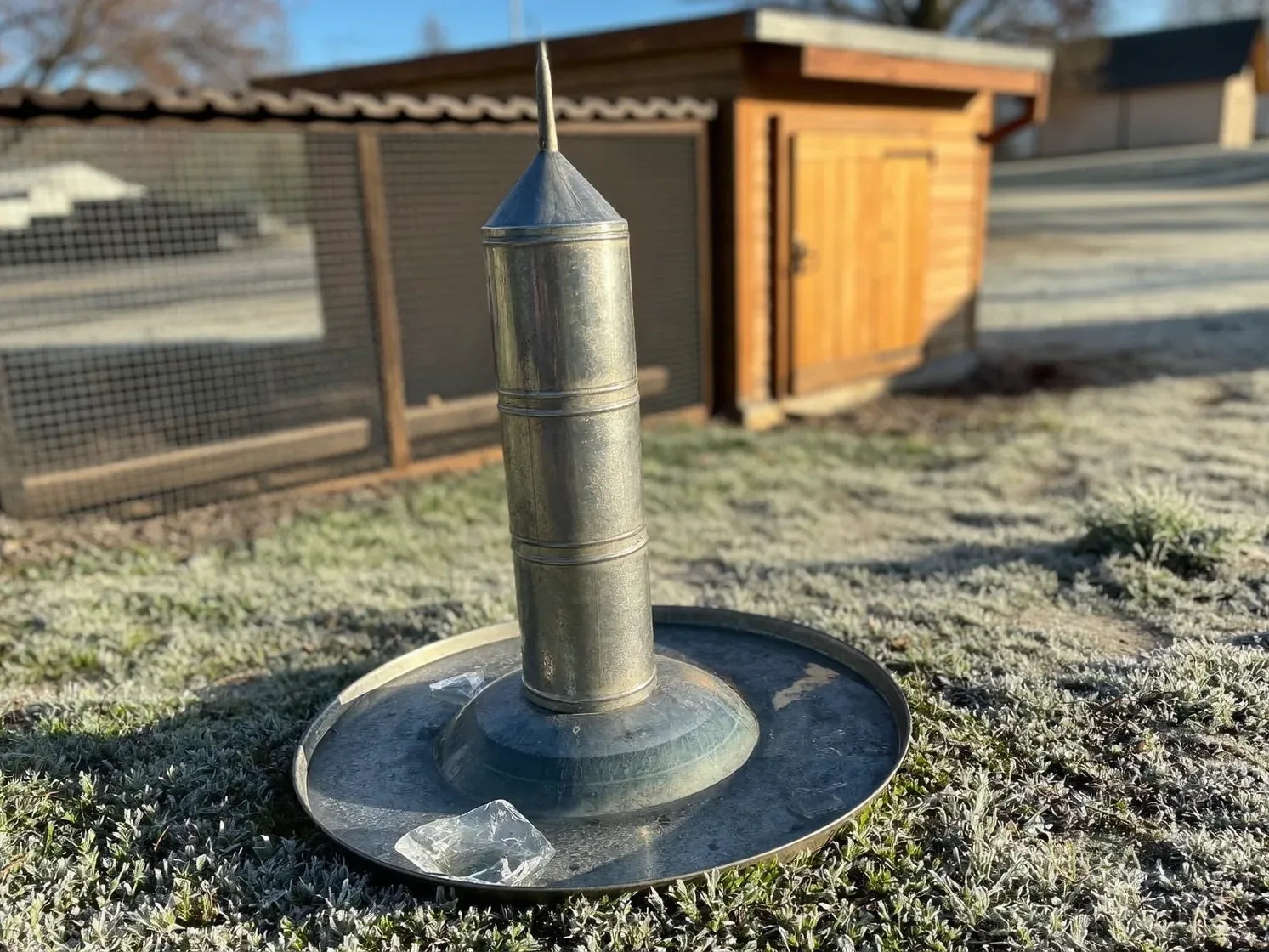 A galvanised metal poultry fount on frosted ground with ice broken in its shallow trough, a wooden coop and run behind